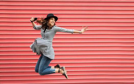 Young woman listening the music and jumping in front of red wall background on the street.