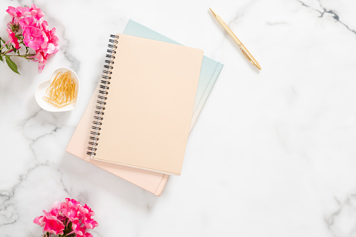 Blank paper notebook, pink flowers, golden stationery on marble background. Flat lay, top view feminine home office desk.