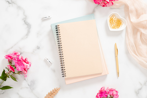 Blank paper notebook, pink flowers, pastel blanket and golden accessories on marble background. Flat lay, top view rose gold home office desk. Beauty blogger workspace concept.