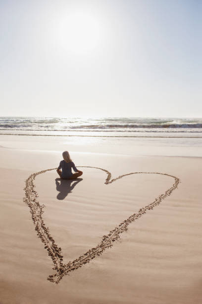 frau sitzt schneidersitz im herzen am strand - beach ideas stock-fotos und bilder