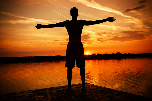 Young man standing with open arms on the pier by the lake.He joying in summer sunset.