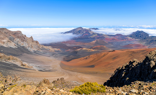 A panoramic view of the colorful volcanic crater at summit of Haleakala, under the bright sun and blue sky, and surrounded by sea of clouds. Maui, Hawaii, USA.