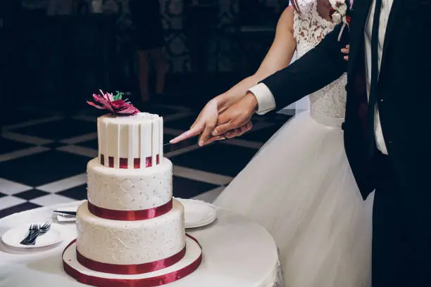 Photo of bride and groom cutting wedding cake. wedding couple tasting big delicious cake with pearl and red flower at wedding reception in restaurant. luxury catering