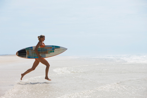 Glad woman in bikini with surfboard smiling and enjoying wind while standing on wet sand near waving sea at sundown on beach