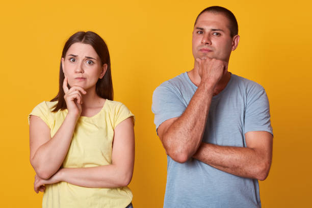 istock Thoughtful beautiful couple looking directly at camera, posing isolated over yellow background in studio, putting hands close to face, keeping in silence, wearing casual tshirts. Togetherness concept. 1163785282