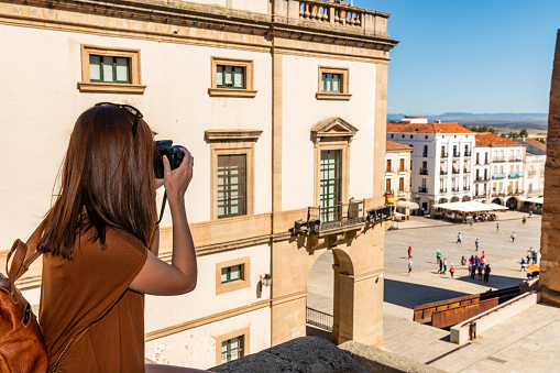 A young red-haired tourist with a backpack takes photographs of the Plaza Mayor in Caceres, Extremadura, Spain.