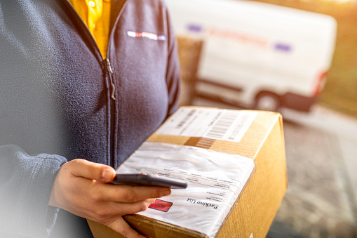 Close up of unrecognisable delivery woman holding in one hand smartphone for signature from home delivery customer and in the other cardboard box, copy space.