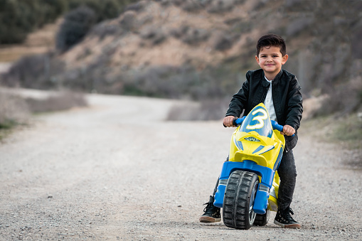 A happy little boy driving a toy motorcycle, dressed in a leather biker jacket on a country road