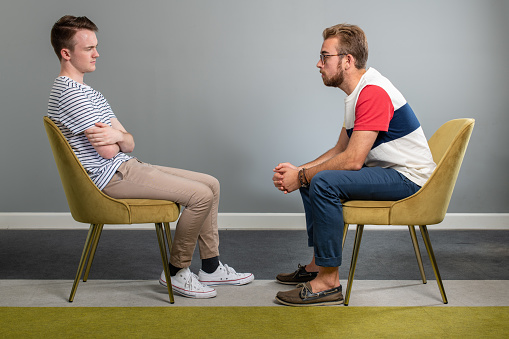 Two Men sit in identical chairs in an empty room while staring directly at each other. One Man leans over as the other sits back in his chair with his arms crossed.