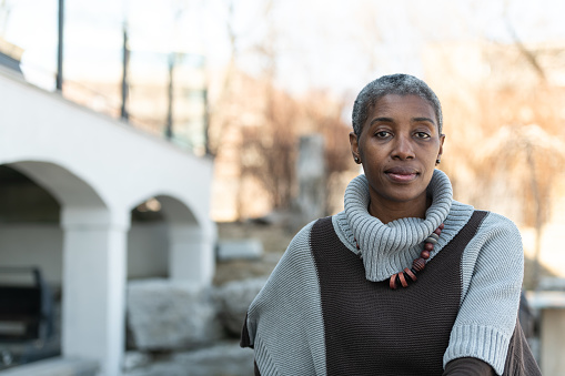 A senior woman of African descent sits outside while looking contemplative. She is dressed warmly in her sweater and looks cozy. She is looking into the camera.
