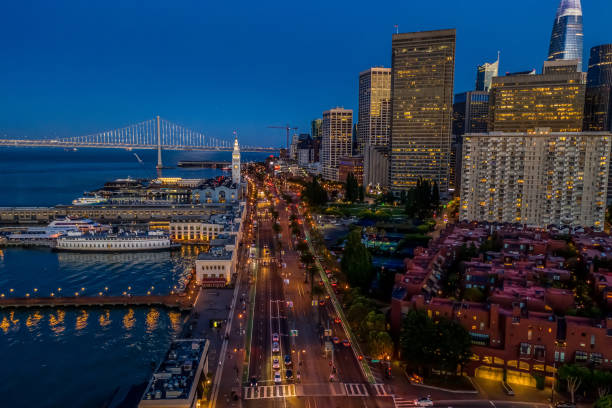 An aerial view of the Ferry Building, Financial District and the Embarcadero at blue hour. City lights twinkle and the illuminated Bay Bridge is seen in the distance.