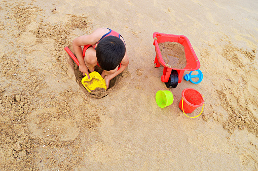 Horizontal photograph of a small child playing with beach toys on sand of a beach in England. The boy in a red vest and shorts is sitting in a small pit and digging sand with a play shovel. A wheelbarrow and bucket and other toys lie along on the sand. Copy space for text.