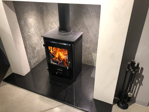 Stock photo of square cast iron woodburner / contemporary log wood burning stove fireplace mantle with orange fire flames burning and generating heat to warm up room instead of gas boiler central heating, modern multifuel stove wood burner stand / chimney flue