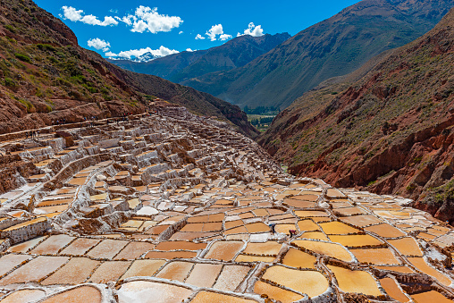 The majestic salt terraces of Maras in the Cusco province with unrecognizable tourists walking between the salt ponds and the snowcapped Salkantay Andes mountain peak in the background, Sacred Valley of the Inca, Peru.