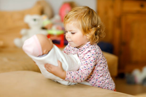 人形で遊ぶ愛らしいかわいい幼児の女の子。幸せな健康な赤ちゃんの子供は、自宅や保育園で母親を再生し、ロールゲームを楽しんでいます。おもちゃでアクティブな娘。 - baby doll dress ストックフォトと画像