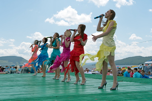 Parnaia, Sharypovskiy district, 
Krasnoyarsk region/ RF - 5 July 2019: Girls in bright dresses sing on a wooden green stage during the ethnic Karatag festival on the shore of a Large lake.