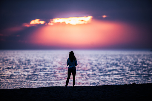 Rear view of a woman standing on the beach and looking at sunset. Copy space.