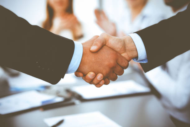 istock Business people shaking hands at meeting while theirs colleagues clapping and applauding. Group of unknown businessmen and women in modern white office. Success teamwork, partnership and handshake concept 1163862837