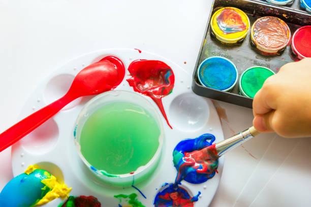 Child's hand holding brush dipped in paint on white paper background with various types gouache watercolor tempera. Kids creativity arts crafts esthetic education. Authentic lifestyle image cepillo de sujeción de la mano del niño sumergido en pintura sobre fondo de papel blanco con varios tipos de temperamento de acuarela gouache. los niños creatividad artes artesanías concepto de educación estética. imagen de estilo de vida auténtica