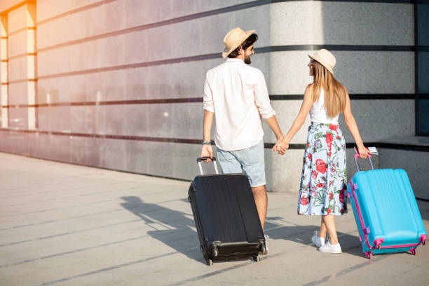 istock Two happy young tourists walking hand in hand away from the camera, pulling suitcases and looking at each other 1163896008