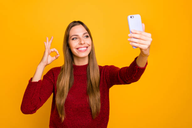 Pretty lady holding telephone hands making selfies showing okey symbol wear knitted pullover isolated yellow background