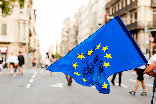 young person walks in the streets in daylight holding european union flag with blood stains on it protesting against immigration policies