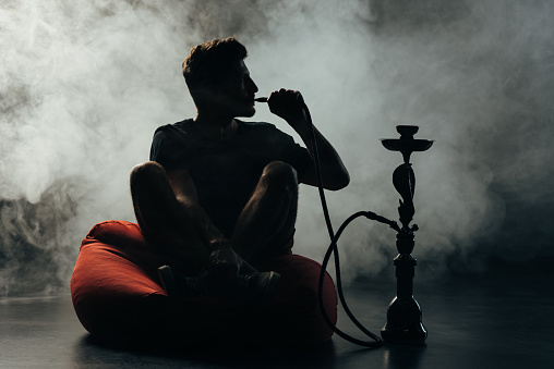 silhouette of man sitting on bean bag chair  with crossed legs and smoking hookah