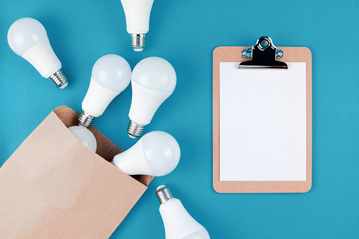 Energy-saving and eco-friendly life in conceptual composition. Creative top view flat lay of LED light bulbs in craft paper shopping bag and clipboard with copy space on blue background minimal style