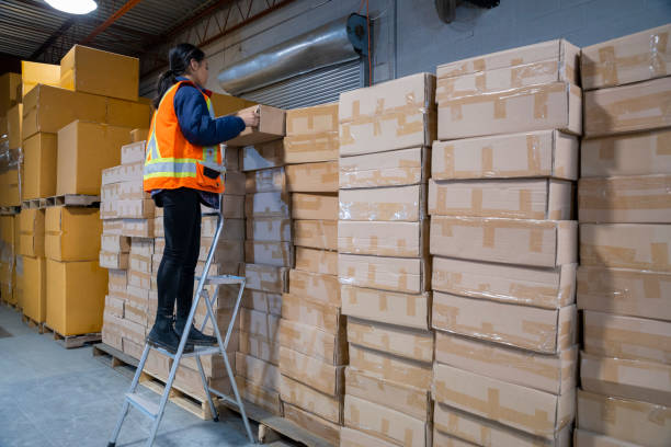 un sujet de sécurité au travail d'entrepôt industriel. une employée utilise une échelle à étapes courtes pour atteindre certaines marchandises placées plus haut. - escabeau photos et images de collection