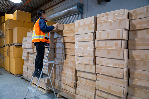 An industrial warehouse workplace safety topic. A female employee uses a short step ladder to reach some higher placed merchandise.