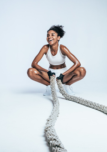 Happy fitness woman taking a break from workout sitting on her toes holding battle ropes. Woman in sportswear relaxing during workout on white background.