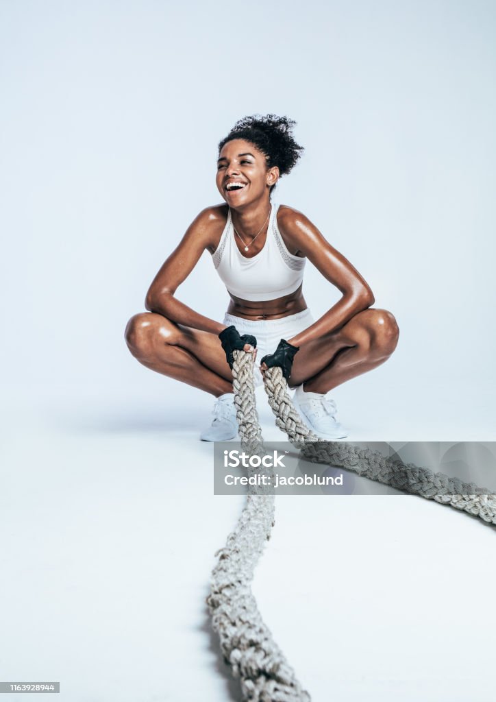 Woman relaxing during workout Happy fitness woman taking a break from workout sitting on her toes holding battle ropes. Woman in sportswear relaxing during workout on white background. Exercising Stock Photo
