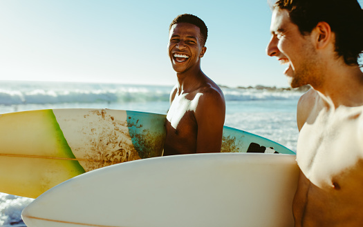 Happy young man with a friend walking on the beach carrying surfboards. Cheerful young friends on vacation at the sea.