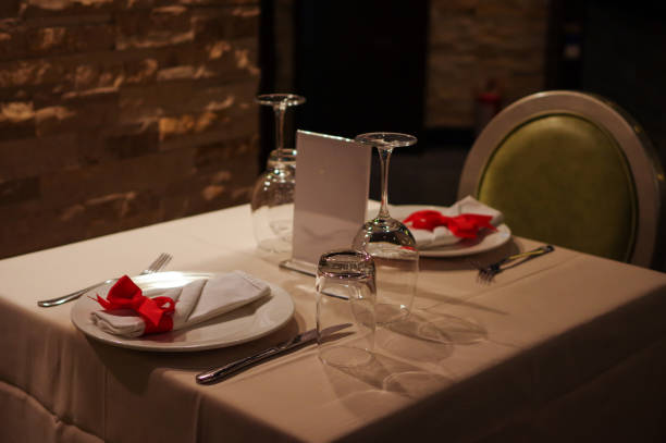 istock Upside down wine glasses and water glasses on restaurant table with knife and fork, napkin decorated with red ribbon on plate and menu holder near brick wall 1163932294