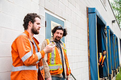 An industrial warehouse workplace safety topic.  Two coworkers smoking a cannabis joint outside of an industrial building while a supervisor or manager secretly looks on.