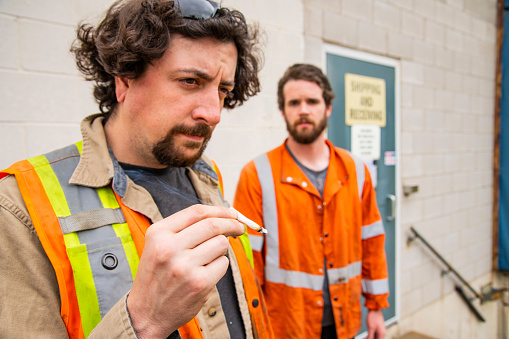 An industrial warehouse workplace safety topic.  Cannabis in the workplace. Two coworkers smoking a cannabis joint outside of an industrial building.