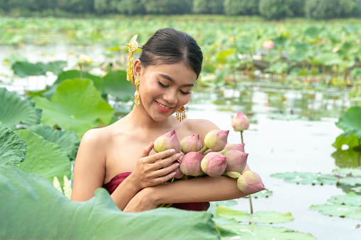 Beautiful asia women wearing traditional Thai dress and sitting on wooden boat in flower lotus lake. Her hands are holding a pink lotus and picking up flower.  And her face is be happy and smile.
