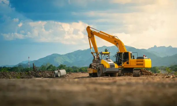 Photo of Backhoe working in road construction site, with mountains and sky background.