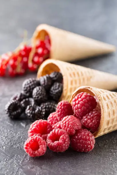 Ice cream cones with berries on a stone background. Summer berries, raspberries, blackberry and currant in waffle cone on dark concrete background. Summer and healthy food concept
