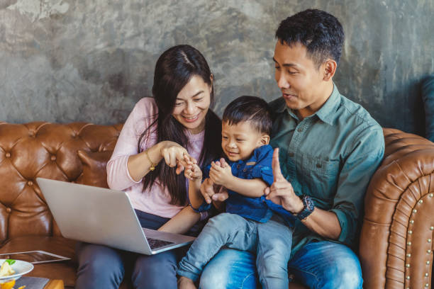 famille asiatique avec le fils regardent le dessin animé par l'intermédiaire de l'ordinateur portatif de technologie et jouant ensemble en vivant dans la maison de grenier pour l'auto-apprentissage ou l'école à la maison, concept d'école à la maison  - brown boy photos et images de collection