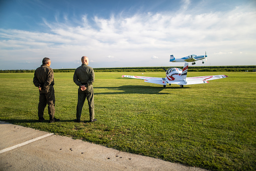 Rear view of two flying instructors, standing and watching small private plane while taking off at the airport