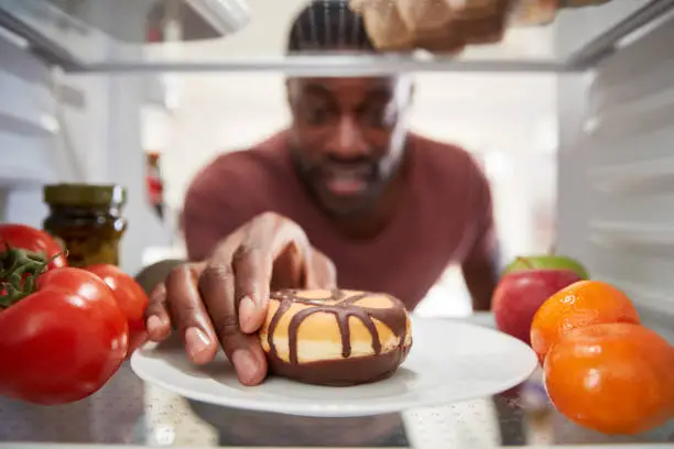 Photo of View Looking Out From Inside Of Refrigerator As Man Opens Door And Reaches For Unhealthy Donut