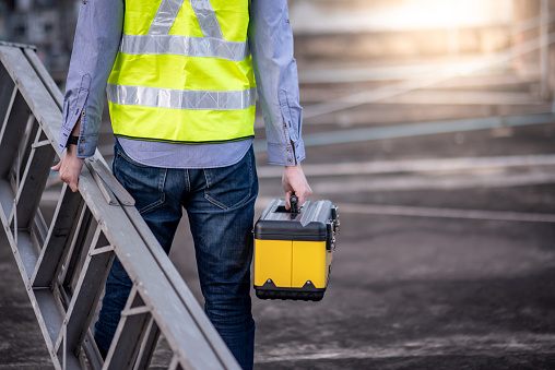 Maintenance worker man with safety helmet and green vest carrying aluminium step ladder and tool box at construction site. Civil engineering, Architecture builder and building service concepts