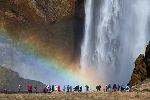Skogafoss, Iceland - April 5, 2018: Waterfall on cliff with rainbow and many tourists people walking with ponchos on trail