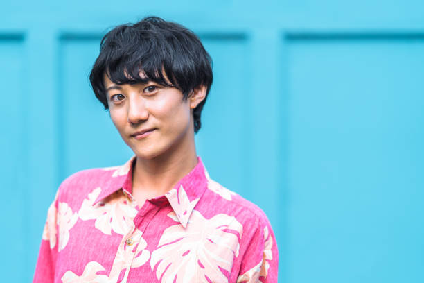 A portrait of a confident modern man wearing floral patterned button down shirt and standing in front of a blue wall.
