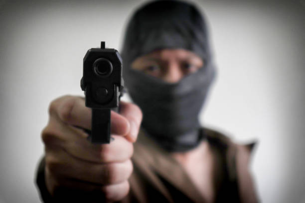 A man is pointing a gun at another man whose face is covered with a cap and with his arms raised.