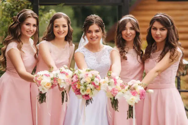 Happy bride with bridesmaid hold bouquets and have fun outside. Beautiful bridesmaid in same dresses stand by the charming bride in long wedding dress.