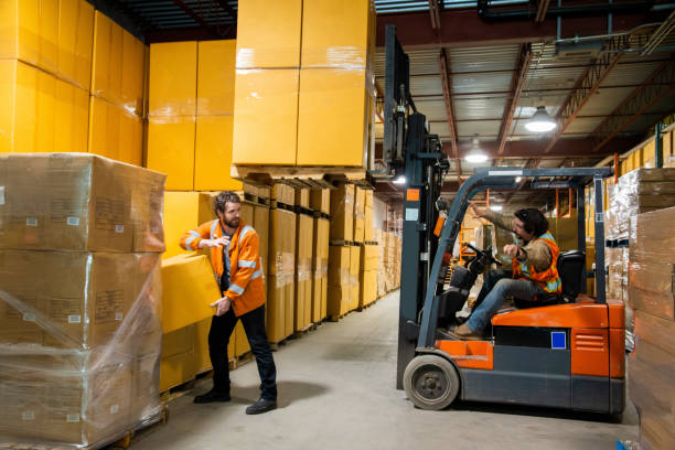An industrial warehouse workplace safety topic. A worker in the danger zone, walking close to a raised forklift carrying a load. An industrial warehouse workplace safety topic. A worker in the danger zone. Injuries caused by forklift accidents are a major safety issue. unsafe workplace stock pictures, royalty-free photos & images