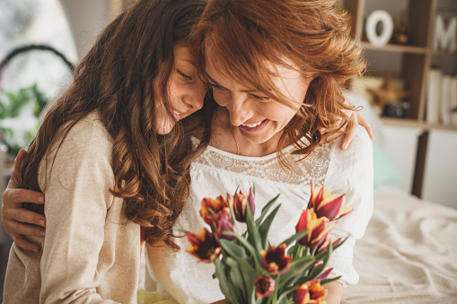 Portrait of a smiling mother and daughter embracing and holding a bouquet of fresh tulips at home.