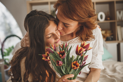 Mother and daughter holding a bouquet of fresh tulips in the living room. Mother is hugging and kissing her daughter.